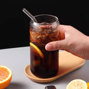 A hand holding a clear glass tumbler with a bent glass straw, filled with iced dark cola and garnished with orange slices, sitting on a wooden coaster with citrus halves nearby.