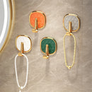 Four wall-mounted jewelry hooks with gold-tone frames and square faceted inserts in white, orange, green, and gray, shown hanging pearl necklaces on a beige marble wall.