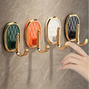 Four wall-mounted hook holders in green, orange, white, and black, each with a gold-tone metal hook and trim, shown mounted on a beige marble-look tiled wall. A hand points toward the rightmost black hook.