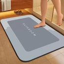 A rectangular bathroom mat with rounded corners in a bathroom setting, featuring a dark gray center with the word 'BATHROOM' in white text. A person's foot is stepping on the mat, and a wooden cabinet, towel, and slippers are visible in the background.