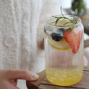A clear glass mason jar filled with a bubbly yellow drink, garnished with a strawberry slice, lemon wheel, blueberry, and rosemary sprig, held on a wooden surface with a hand visible at the left edge.