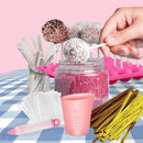 A hand is dipping a white chocolate ball into pink sprinkles in a clear jar, surrounded by candy-making accessories including gold and white sticks, a pink mold tray, a pink measuring cup, and white wrappers on a blue-and-white checkered tablecloth against a pink background.