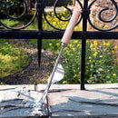 A garden weeding tool with a light wood handle and stainless steel forked prongs, shown resting against a black wrought iron fence on a stone ledge with a garden background.