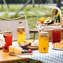 Outdoor picnic scene with four glass mason jar drinks with bamboo lids and metal straws, set on a table with pastries, fruit, and a wicker basket in a blurred garden background.