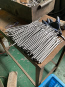 A pile of long silver metal rods or prongs spread across a worn wooden workbench, with a blue work glove and safety glasses nearby in an industrial workshop setting.