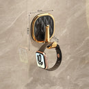 Gold oval wall hook with a black faceted center and a smartwatch hanging from it, shown on a beige marble-look wall with dimension labels.