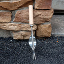 A metal garden spade fork with a light wood handle, shown standing upright against a stone wall on a bed of dark gravel.